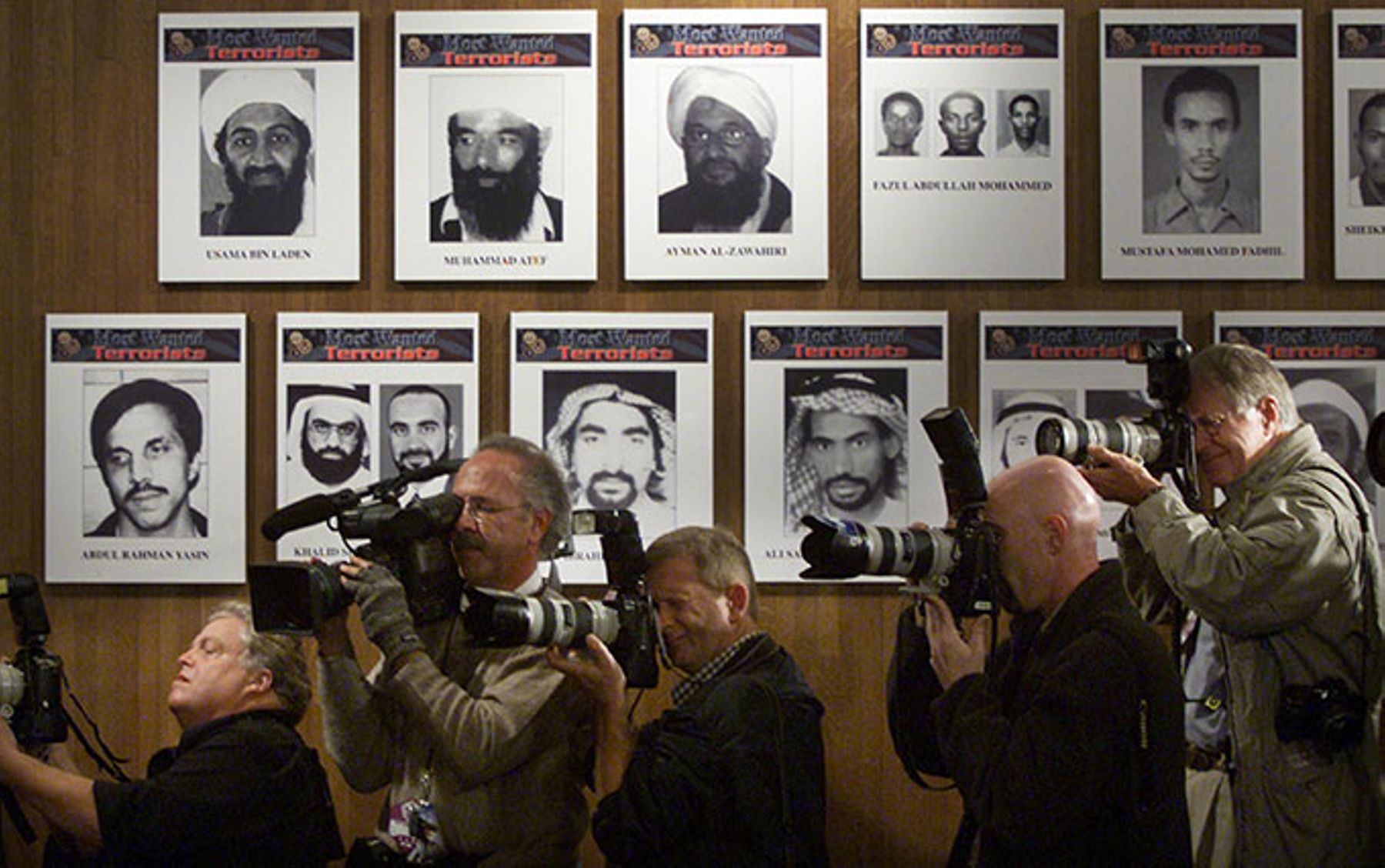 Members of the media stand next to a wall of the Most Wanted terrorists.