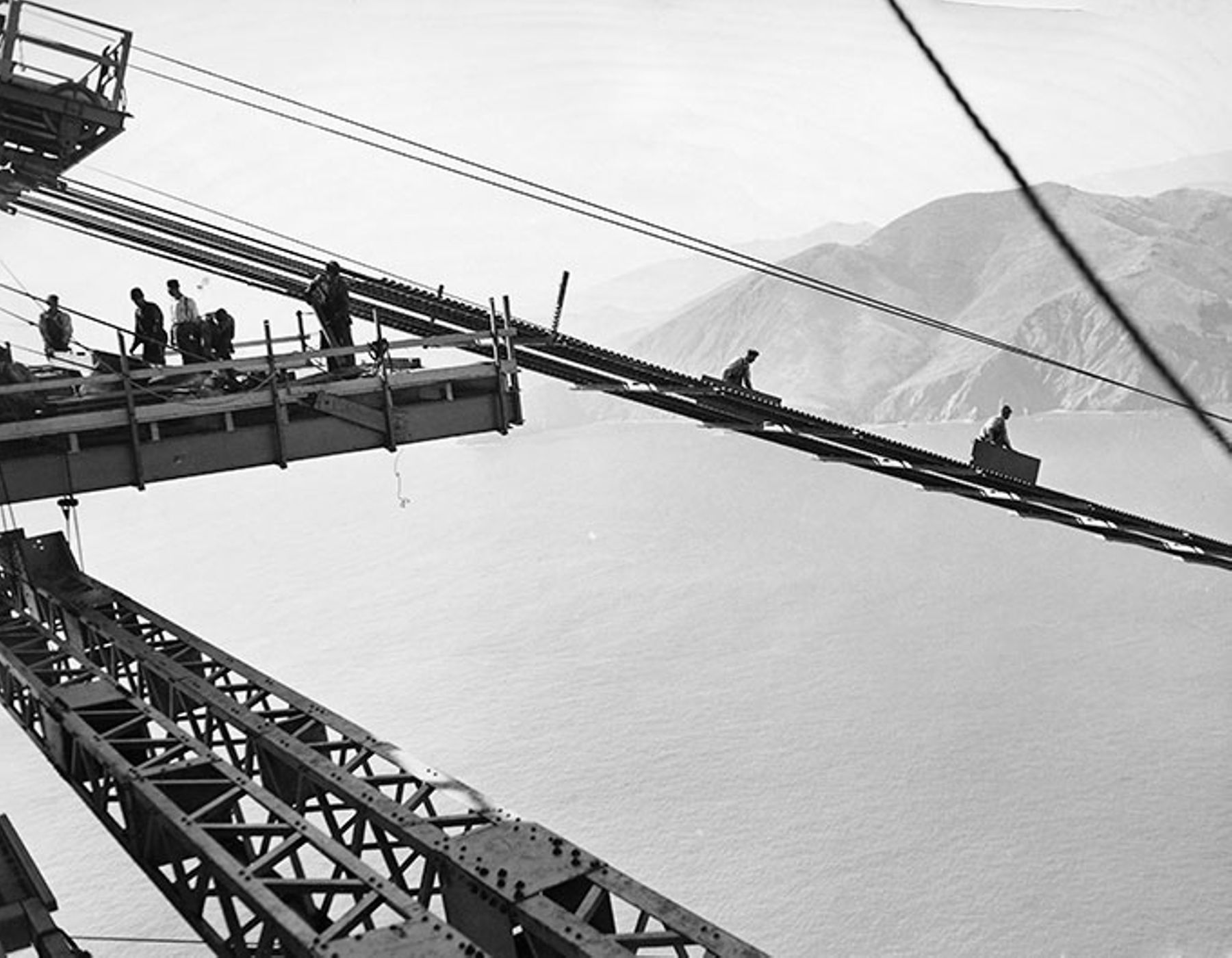 The Golden Gate Bridge under construction. men are working on the catwalk, sending pieces down a ramp system.